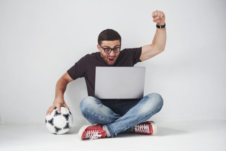 Excited soccer fan with a football isolated on white background. He sits on the floor and watches the results of the game and the best moments of the match on the laptop